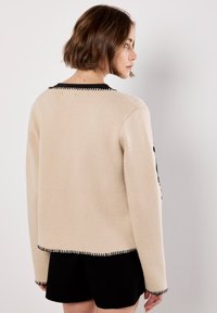 Beige Strickpullover mit langen Ärmeln, schwarzem Besatz am Kragen und Saum, verziert mit dekorativen schwarzen Nähten entlang der Ränder.