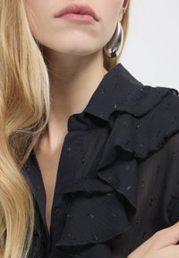 Donna bionda con capelli lunghi che indossa un grande orecchino a cerchio in argento e una blusa nera con texture, colletto a volant e dettagli di paillettes sottili.