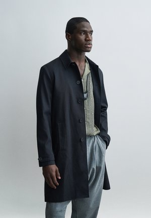 Homme portant un manteau noir boutonné sur une chemise à motifs et un pantalon gris, debout avec une main dans la poche, sur un fond clair uni.
