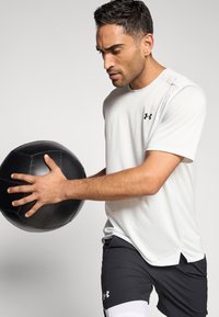 Camiseta deportiva blanca de manga corta, con logo negro, y pantalones cortos negros con detalles blancos; sosteniendo una pelota medicinal negra texturizada.