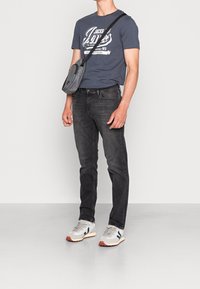 Vaqueros gris oscuro, camiseta azul marino con estampado gráfico blanco y bolso bandolera gris. Las zapatillas deportivas son gris y blanco con detalles en naranja.