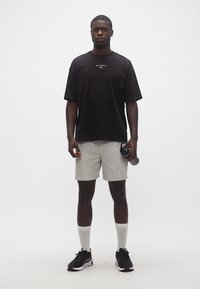 Άνδρας που φοράει μαύρο t-shirt Puma Hyrox, γκρι σορτς, λευκές κάλτσες και μαύρα αθλητικά παπούτσια, κρατώντας μια μαύρη φιάλη νερού σε λευκό φόντο.