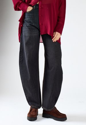 Person iført løse sorte jeans, en rød skjorte med knapper og brune snørebinding sko står på en hvid baggrund.