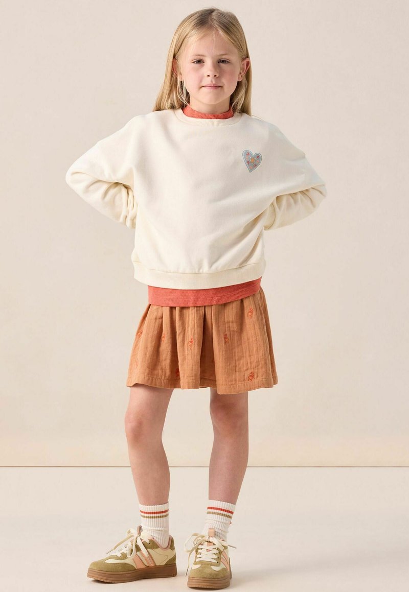 Sweatshirt crème avec bordures orange, patch en forme de cœur ; jupe couleur rouille avec broderies florales ; chaussettes blanches avec rayures rouges ; baskets vertes et beiges.