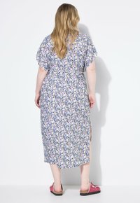 Abito maxi floreale in viola, blu e giallo, con maniche corte e aperture laterali, abbinato a scarpe slip-on rosa su sfondo bianco.