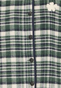 Chemise en flanelle à carreaux verts et navy, ornée d'une rangée de boutons noirs et d'un détail brodé floral blanc. Texture douce avec une finition contrastante.