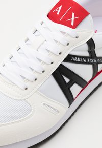Sneakers bianchi con accenti neri e rossi, caratterizzati da una parte superiore in pelle liscia, suola in gomma e un logo "A/X" prominente sulla linguetta.