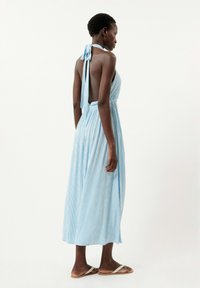 Hellblaues gestreiftes Halterkleid mit offenem Rücken und gebundenem Hals. Fließender Stoff, der bis zu den Knöcheln fällt. Trägt flache Sandalen.