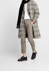 Person med svart scarf, lång grå rutig kappa, beige rutiga byxor, vit skjorta och svarta läderskor som står med en hand i fickan.