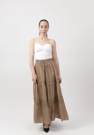 Frau mit dunklen Haaren, die ein weißes ärmelloses Oberteil und einen langen beigen Rock mit Spitzenverzierung trägt, stehend vor einem schlichten hellen Hintergrund.