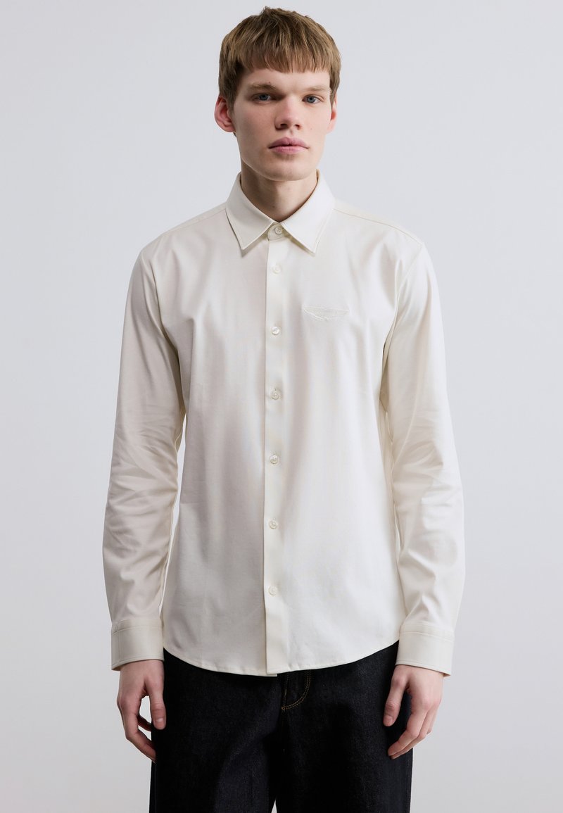 Κρεμ πουκάμισο με κουμπιά, γιακά διασποράς, μακριά μανίκια και διακριτικό κεντητό σχέδιο στο στήθος. Απαλό ύφασμα, λεία υφή.