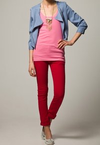 Lichtblauwe cropped jas, roze tanktop, rode skinny jeans en zilveren instappers. Halsketting met gouden en roze accenten. Effen achtergrond.