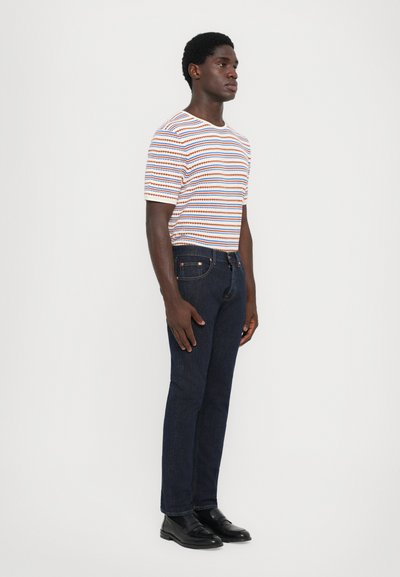 Camiseta de rayas en blanco, naranja y azul; jeans de mezclilla azul oscuro; zapatos negros. El modelo está de lado contra un fondo liso.