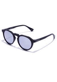 Črne osmerokotne sončne očala z obarvanimi lečami, debelimi okvirji in elegantnimi kraki. Izdelano v Španiji, na strani je znamka "Hawkers".