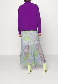 Lila Langarmshirt kombiniert mit einem fließenden, mehrfarbigen Rock mit einem grün- und lila-gepunkteten Muster. Helle grüne Stiefeletten.