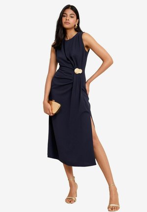 Navy blue sleeveless dress with twisted front detail, side slit, and circular gold hardware. Complemented by a gold clutch and strappy heels.