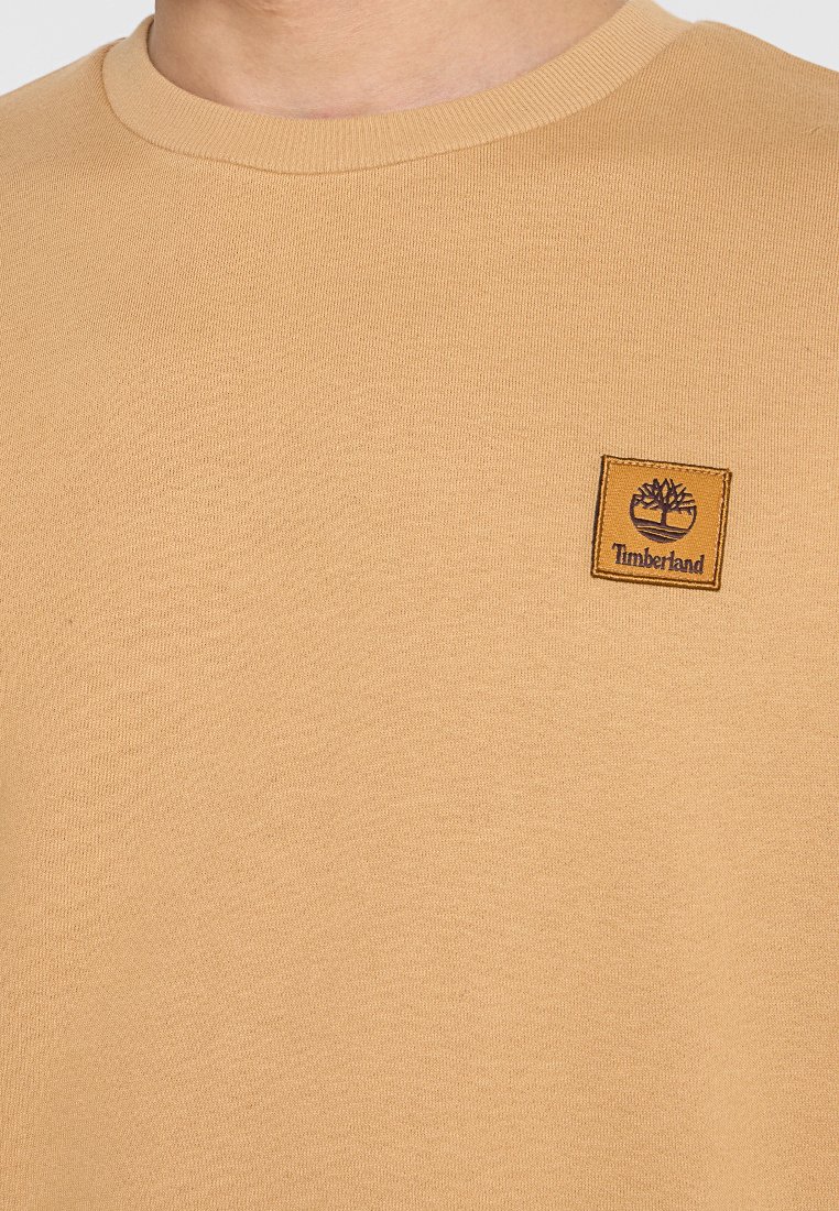 T-shirt beige a maniche corte realizzato in tessuto testurizzato, con un patch del logo quadrato Timberland sul lato sinistro del petto. Collo rotondo.