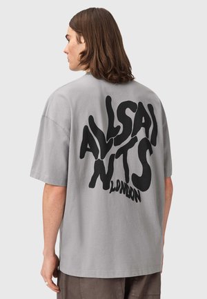 Persoon met halflang bruin haar die een grijs oversized T-shirt draagt met grote zwarte tekst "AllSaints London" op de achterkant.