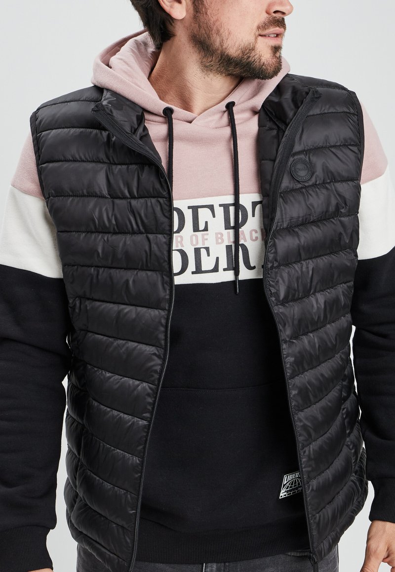 Chaleco acolchado negro con textura acolchada, cierre de cremallera, usado sobre una sudadera negra y rosa con un diseño gráfico y capucha con cordón.