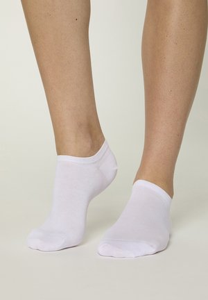 Nohy v obyčejných bílých kotníkových ponožkách stojící na světlém neutrálním povrchu a pozadí.