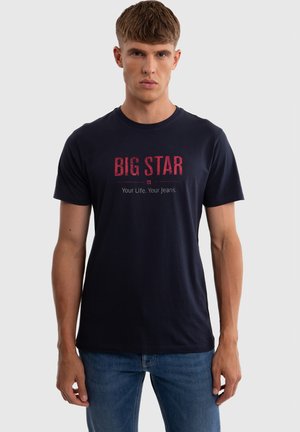 Noor mees, kes kannab tumesinist T-särki kirjaga "BIG STAR Your Life. Your Jeans." ja siniseid teksaseid, seisab ühevärvilise hele tausta ees.