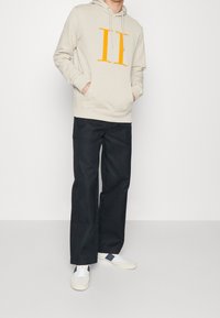 Sudadera beige con un gran gráfico naranja "II", combinada con pantalones oscuros holgados y zapatillas blancas con acentos en azul marino.