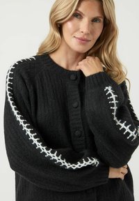 Svart stickad cardigan med en strukturerad design, rund halsringning och stora knappar. Vita stickade detaljer på ärmarna ger extra finish.