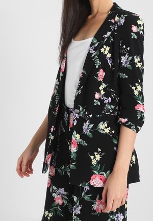 Kvinde iført en sort blomsterblazer med 3/4-ærmer og matchende bukser over en hvid top, poserer mod en ensfarvet lys baggrund.
