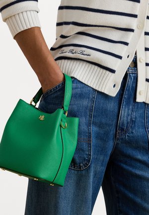 Žalias odinis kibirinis krepšys su auksiniais aksesuarais, laikomas prieš mėlynus džinsus, su balta ir tamsiai mėlyna juostuota kardigana.