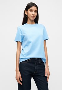 Γυναίκα με μακριά σκούρα μαλλιά φορώντας χρυσούς κρίκους σκουλαρίκια, ένα ανοιχτό μπλε Τ-shirt και σκούρα μπλε τζιν πάνω σε ένα απλό ανοιχτό φόντο.