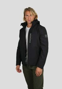 Homme blond portant une veste noire zippée avec capuche, une chemise blanche en dessous, et un pantalon vert olive, se tenant contre un fond gris.