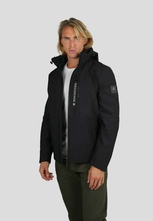 Blonde man in een zwarte ritsjack met capuchon, een wit shirt eronder en olijfgroene broek, staand tegen een grijze achtergrond.