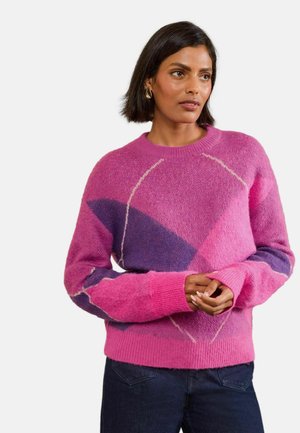 Mujer con el cabello corto y oscuro que lleva pendientes de aro dorados y un suéter de punto con estampado rosa y púrpura, de pie con las manos ligeramente entrelazadas.