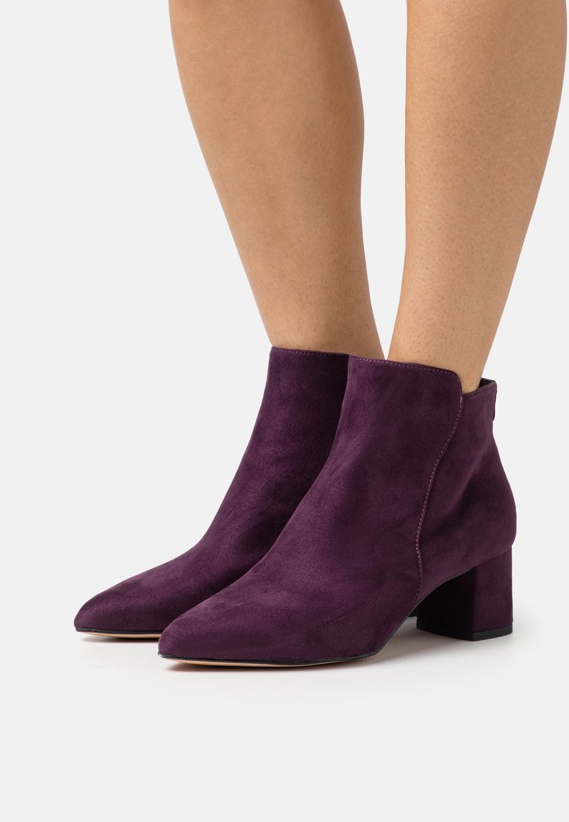 Jambes portant des bottines en daim violettes à bouts pointus et talons carrés sur un fond blanc uni.