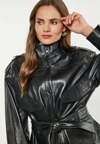 Giubbotto in pelle nera con colletto alto, chiusura frontale a bottoni e ampie tasche al petto. Include una vita con cintura per una vestibilità strutturata.