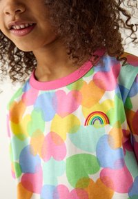 T-shirt di cotone colorata con grandi cuori in rosa, verde, blu, arancione e giallo, con un piccolo arcobaleno ricamato sul petto.