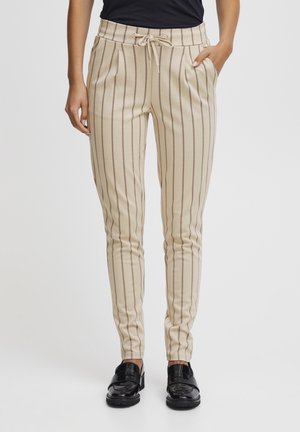 Slim-Fit beige Hose mit vertikalen braunen Streifen, elastischem Bund mit Kordelzug, Seitentaschen, getragen mit schwarzen Schuhen und dunklem Oberteil.