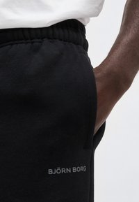 Κοντινό πλάνο ενός ατόμου που φοράει μαύρο παντελόνι φόρμας Björn Borg με ελαστική μέση και το χέρι του να ακουμπά στην τσέπη.