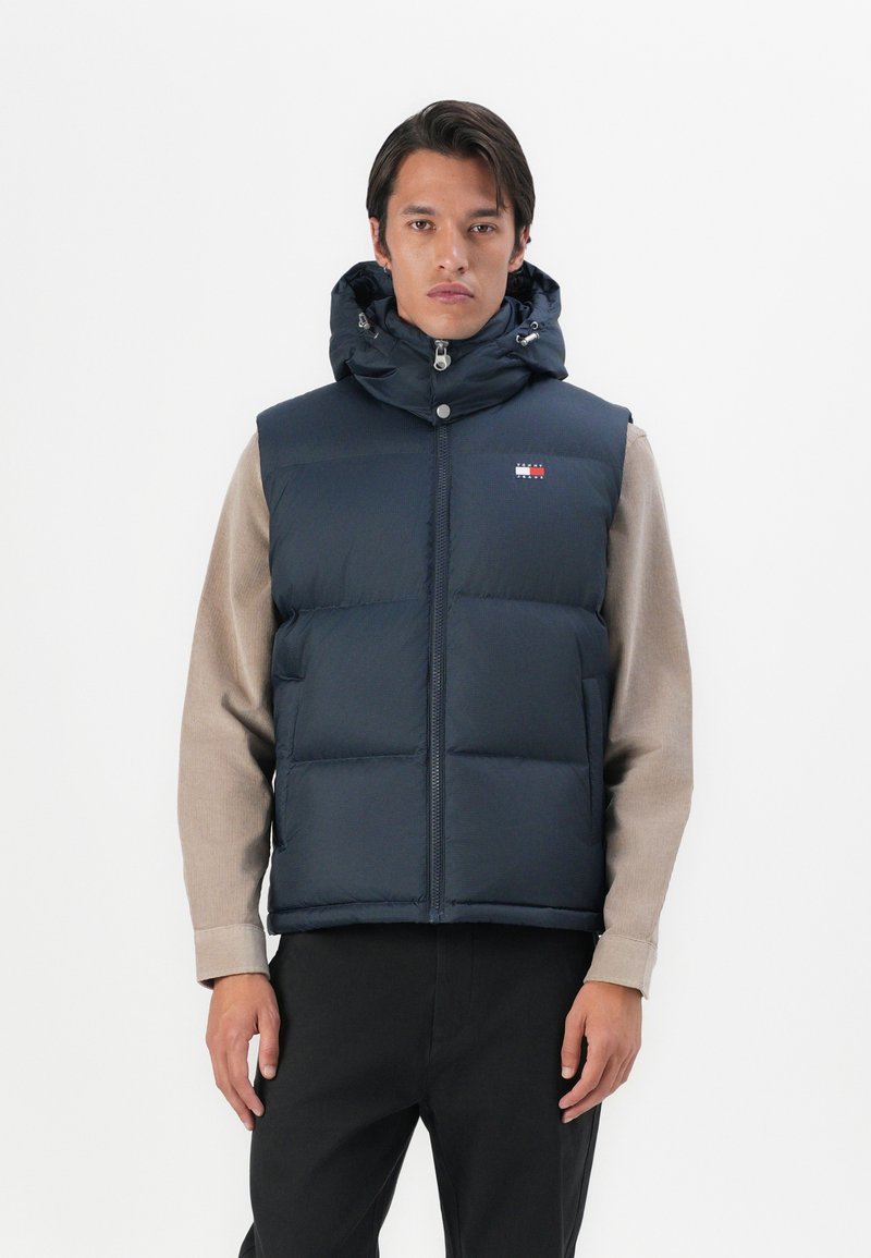 Navy blue padded vest with a hood, zip closure, front pockets, and a logo patch. Fabric appears smooth and slightly glossy.