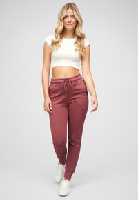 Bordeauxfarbene Sweatpants aus weichem Stoff mit elastischem Bund und seitlichen Taschen, kombiniert mit einem weißen, kurzen Oberteil und weißen Sneakers.