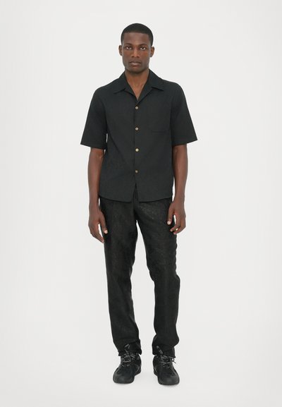 Chemise noire à manches courtes avec des boutons dorés et une poche, associée à un pantalon noir à motifs et des baskets noires.