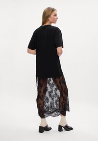Kobieta stoi bokiem, ubrana w czarny, oversize'owy T-shirt i przezroczystą czarną koronkową spódnicę, a na nogach ma szpilki i jasne skarpetki.