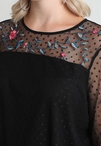 Blouse noire avec une superposition transparente à pois et des broderies florales colorées sur les épaules. Présente un col rond et des manches courtes.