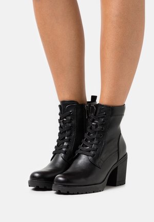 Bottes en cuir noir montant jusqu'au milieu du mollet, lacées avec une fermeture éclair latérale et des talons épais, portées sur des jambes bronzées, sur fond blanc.