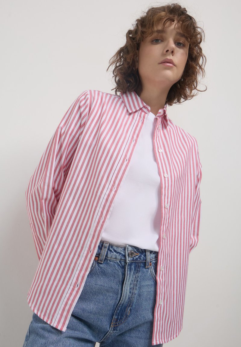 Junge Person mit lockigem Haar, die ein rosa-weiß gestreiftes Hemd über einem weißen Oberteil und blauen Jeans trägt, vor einem schlichten Hintergrund.