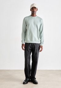 Man staat in een lichtgrijze sweatshirt, beige pet, zwarte spijkerbroek en zwarte loafers tegen een witte achtergrond.