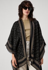 Zwarte sjaal met gouden accenten, met een herhalend tekstpatroon, franjes aan de randen en een open voorkant. Gedragen over een beige top.