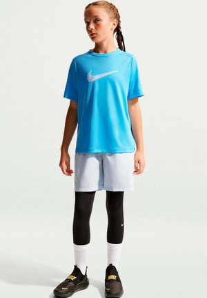 Fille aux cheveux tressés portant un t-shirt bleu Nike, un short clair, des leggings noirs, des chaussettes blanches et des baskets noires Nike, debout devant un fond uni.
