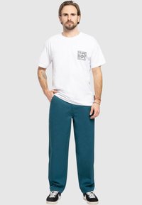 T-shirt bianca a maniche corte con un design grafico sulla tasca, abbinata a pantaloni della tuta color teal. Presenta sneaker nere con suole e lacci bianchi.