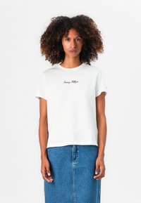 T-shirt en coton blanc à manches courtes et col rond, avec logo "Tommy Hilfiger" brodé en bordeaux. Associé à une jupe en denim.